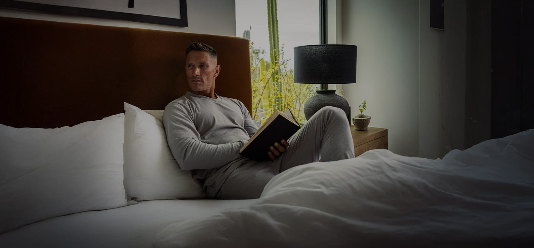 A man in light gray loungewear sits on a bed holding an open book, looking to the side. There is a lamp and a small plant on the bedside table near a window.