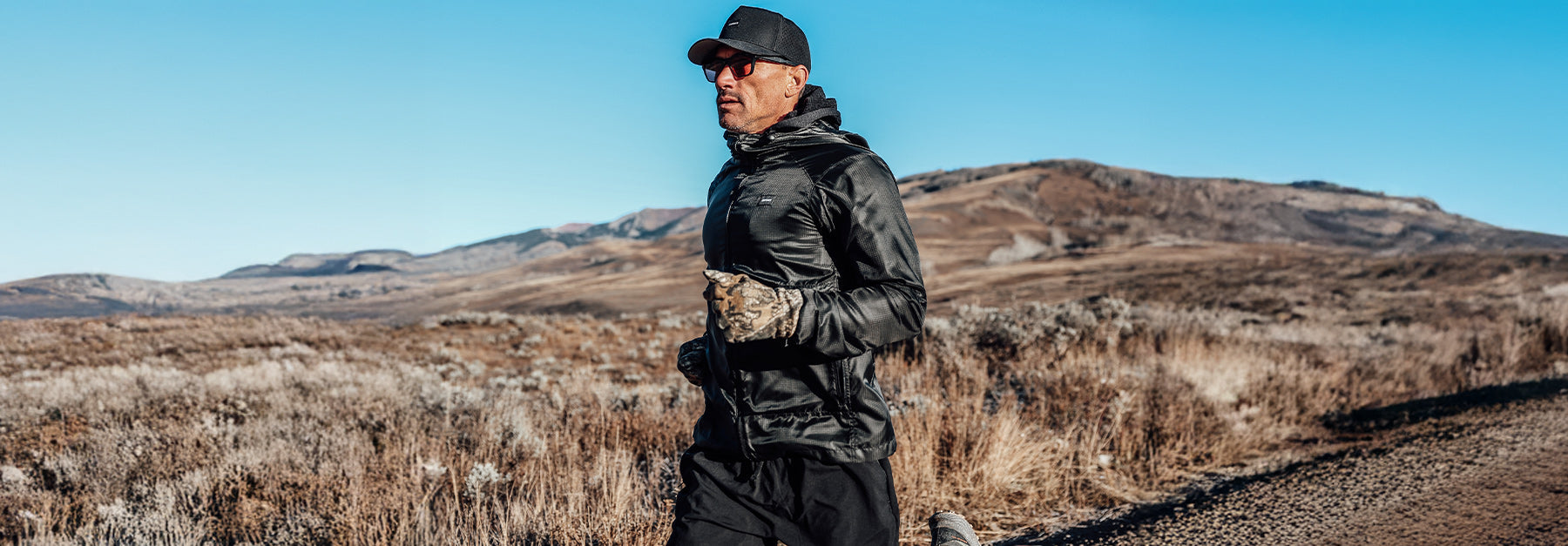 A man wearing sunglasses, a black jacket, gloves, and a cap is running on a dirt path through a dry, open landscape with hills in the background under a clear blue sky.