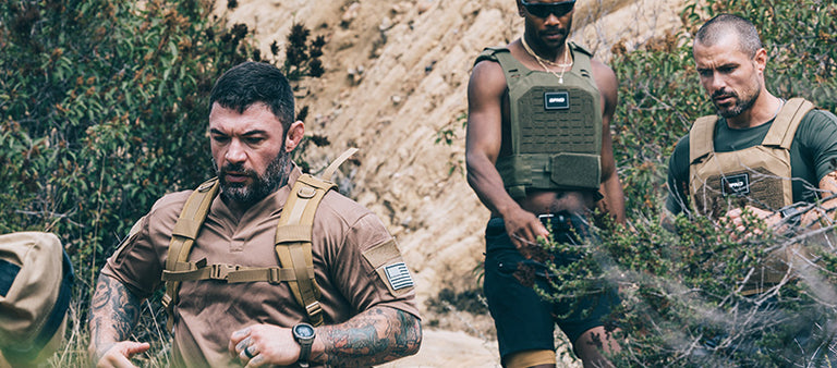 Three men wearing tactical gear and plate carriers move through a rugged outdoor environment with shrubs and rocks.