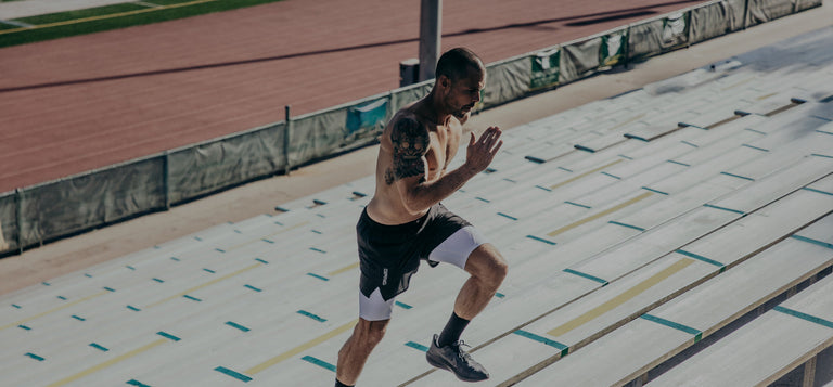 Shirtless man with tattoos runs up stadium bleacher steps during daytime, wearing athletic shorts and shoes.