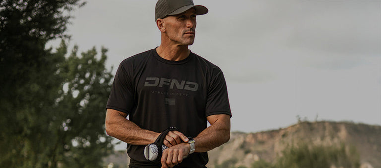 Man wearing a black DFND t-shirt and cap stands outdoors, holding a black leash or strap, with trees and hills in the background.