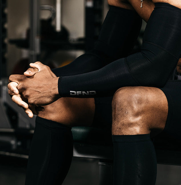 A person wearing black compression sleeves sits on a gym bench with hands clasped, surrounded by gym equipment and weights in the background.