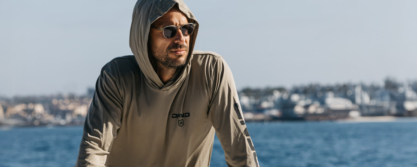 A person wearing sunglasses and a hooded tan long-sleeve shirt stands outdoors near water with a blurred cityscape in the background.