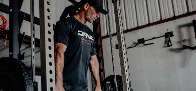 Man wearing a black t-shirt and cap standing in a gym, gripping weights, with gym equipment and wall-mounted bars in the background.