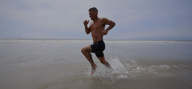 A shirtless man in black shorts runs through shallow water at the beach under a cloudy sky.