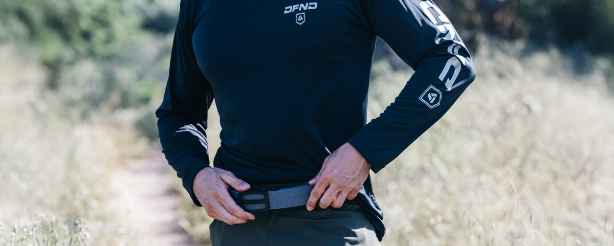 Person outdoors in a long-sleeve black DFND shirt adjusting a belt, standing on a dirt path surrounded by tall grass and greenery.