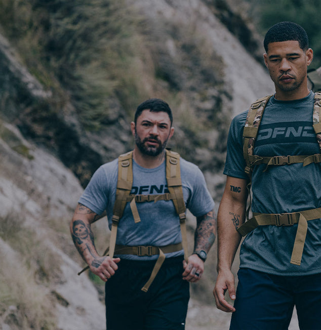 A man wearing a cap, black shirt, shorts, and a backpack runs on a dirt trail between rocky cliffs and greenery.