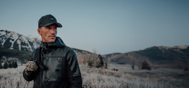 A man in a dark jacket and cap stands outdoors in a frosty landscape with mountains and trees in the background.