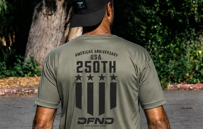 A person wearing an olive green shirt with "Americas Anniversary USA 250th" and black graphic designs on the back, standing outdoors.