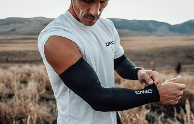 Person standing in a field wearing black compression leggings under black shorts, a white shirt, and white socks. Their legs and arms have visible tattoos.
