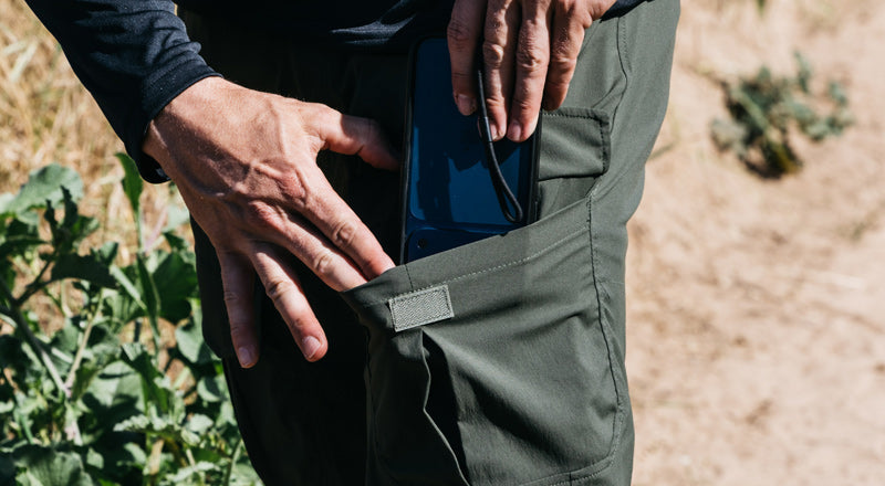 A person puts a black smartphone into the side pocket of green cargo pants while standing outdoors on a dirt path near green plants.