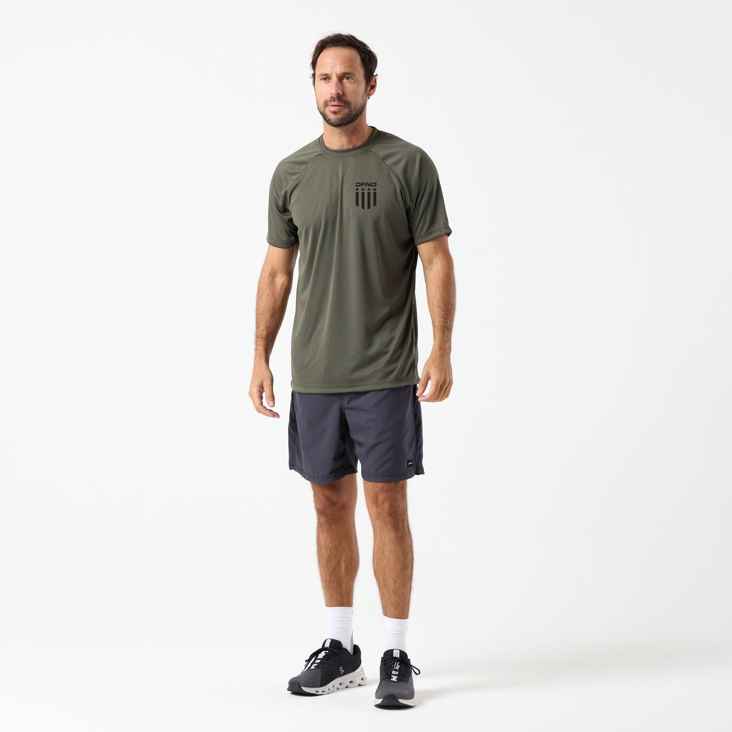 A man faces forward against a white background wearing a DFND SPEED TECH 250TH SS SHIRT in olive green, dark gray shorts, white socks, and black athletic shoes.