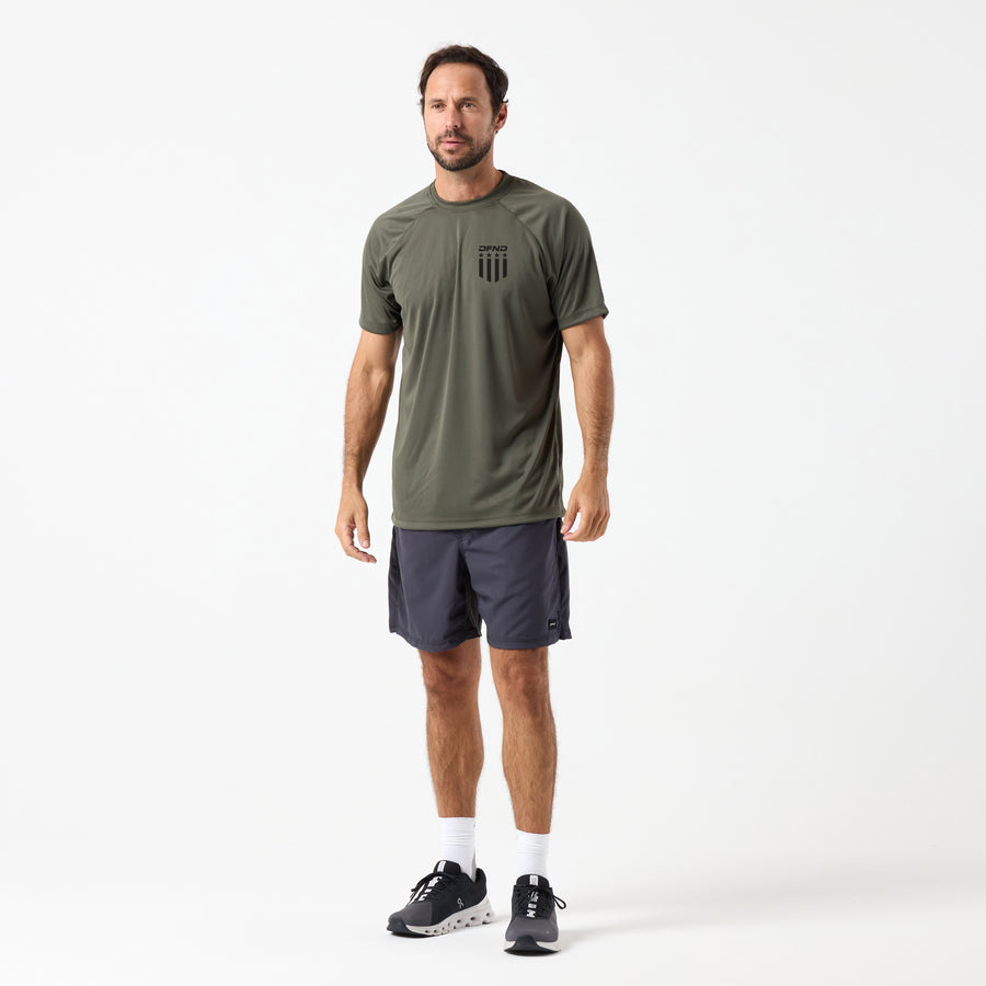 A man faces forward against a white background wearing a DFND SPEED TECH 250TH SS SHIRT in olive green, dark gray shorts, white socks, and black athletic shoes.