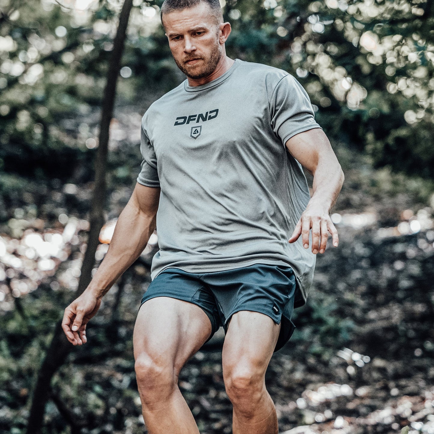A man wearing the DFND Speed Tech Short Sleeve + Aero Short Outdoor Kit is walking or running outdoors in a forested area, enjoying its quick-drying fabric for comfort.
