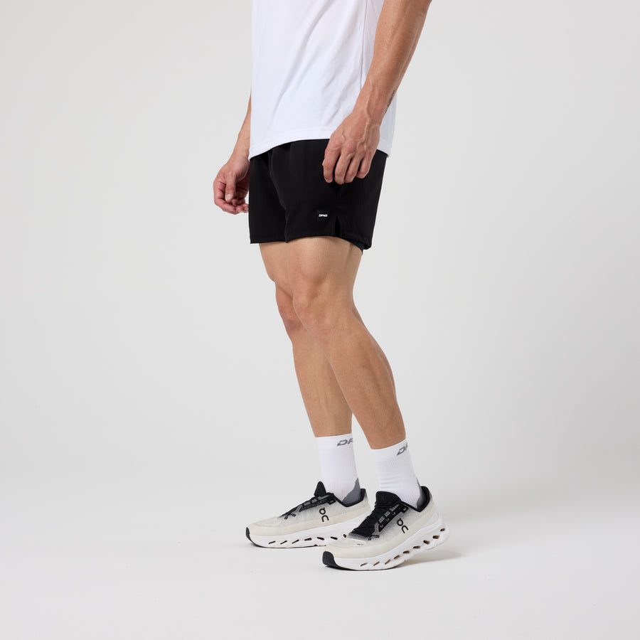 Person standing against a plain background wearing a white shirt, DFND AERO 2N1 SHORT 5" compression training shorts made in USA, white socks, and white athletic shoes with black details.