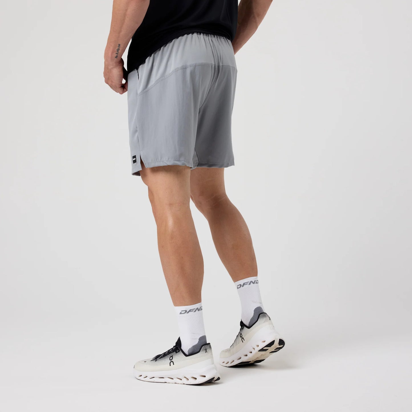 A man stands against a plain background wearing the DFND Speed Tech Short Sleeve + Aero Short Outdoor Kit, featuring a black shirt, light gray quick-drying athletic shorts, white socks, and white athletic shoes.