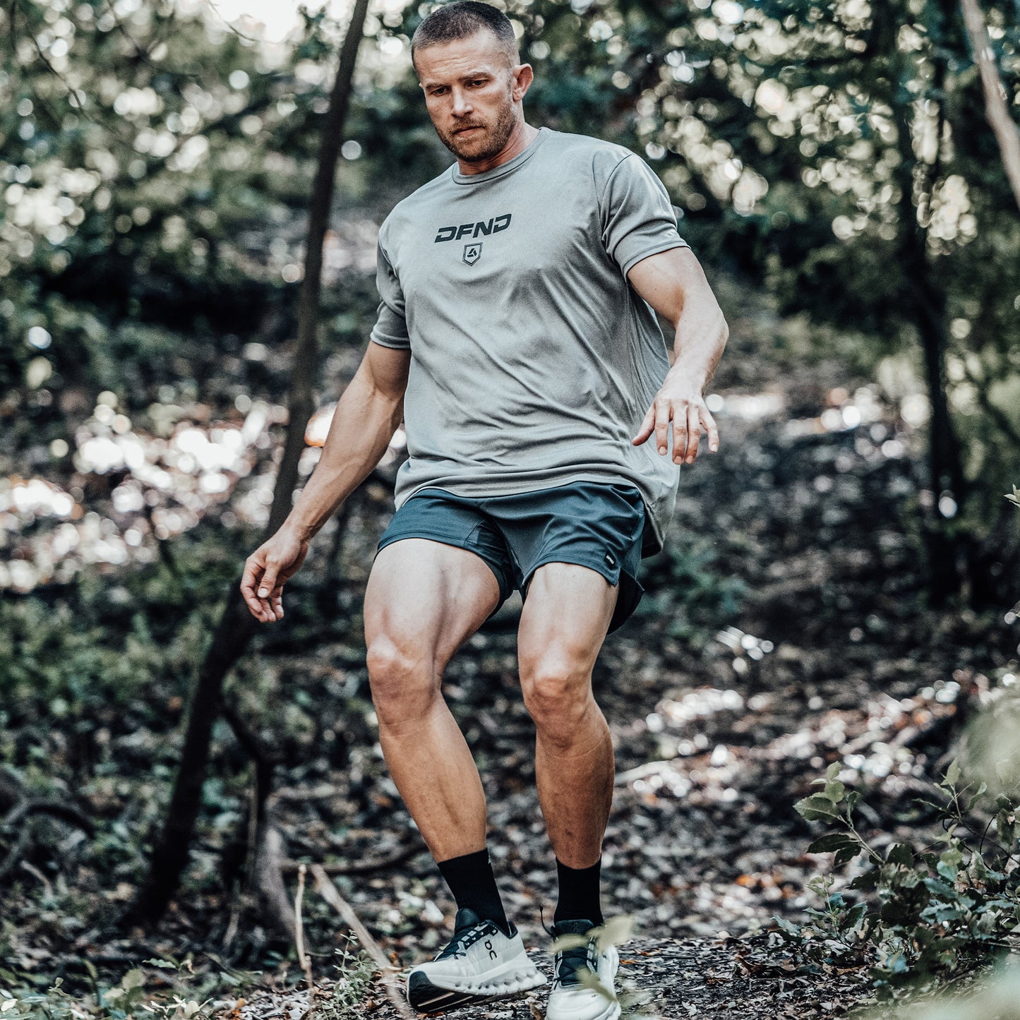 A man wearing athletic clothing, including DFND AERO SHORT 5″ performance training shorts made in the USA, walks downhill on a dirt trail through a wooded area, surrounded by trees and greenery.