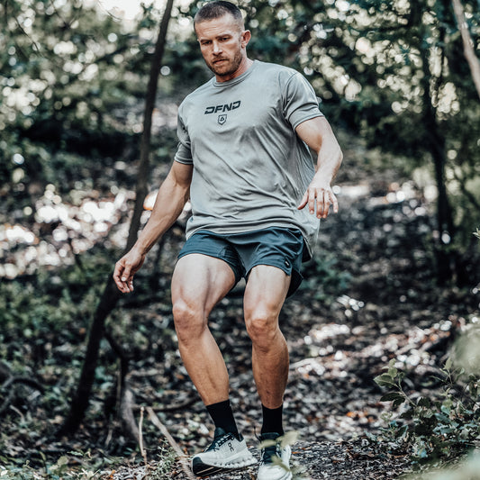 A man wearing athletic clothing, including DFND AERO SHORT 5″ performance training shorts made in the USA, walks downhill on a dirt trail through a wooded area, surrounded by trees and greenery.