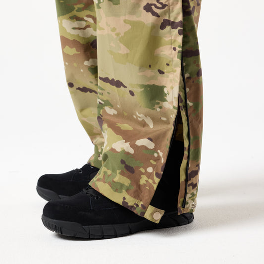 A person wearing DFND Assault Pants with snap buttons on the sides and black shoes stands on a white surface.