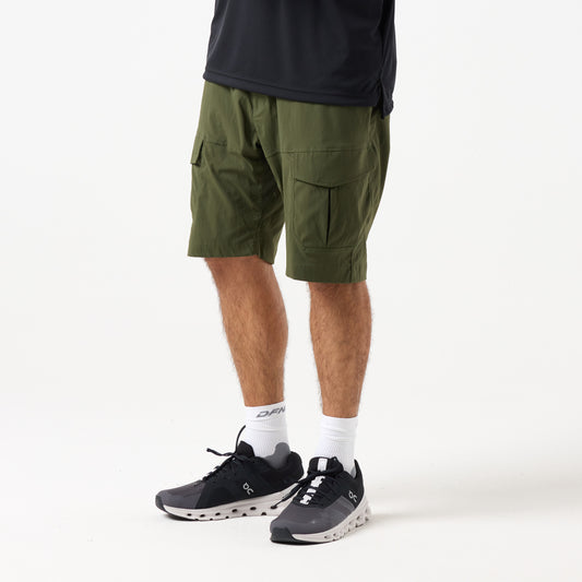A person stands against a plain white background wearing DFND's MISSION CARGO SHORTS in olive green, featuring zippered cargo pockets, paired with a black shirt, white crew socks, and black and white sneakers.