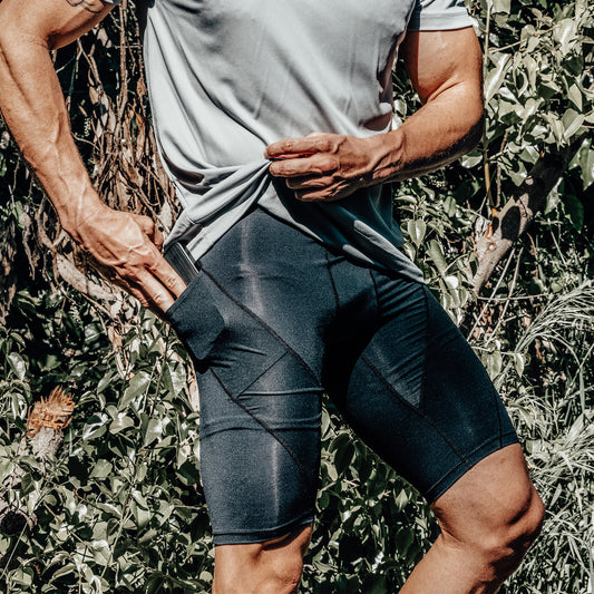 While outdoors, a person tugs at the pocket of their DFND Active Ax Compression Shorts, highlighting the functional pocket detail and emphasizing both contemporary style and reliable pelvic stability.