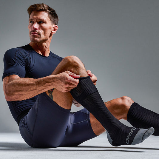 A man in athletic wear sits on the floor against a plain background, adjusting his DFND HYBRID Ax / Rx COMPRESSION SOCKS 20-30mmHg while looking to the side.