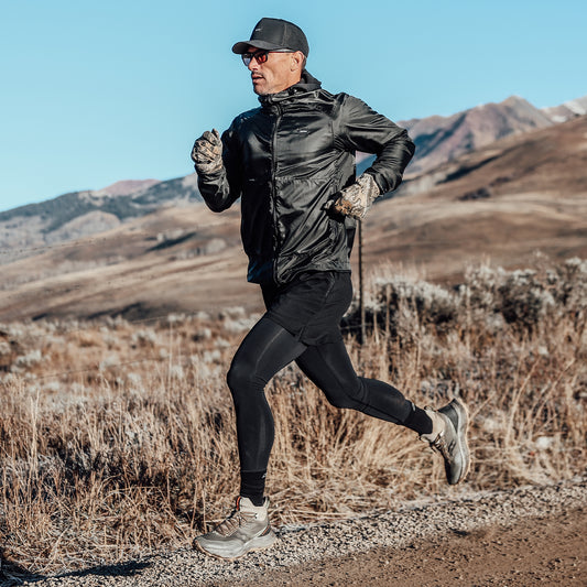 Wearing DFND’s RECOVER Rx COMPRESSION TIGHT, a man runs outdoors on a gravel path through a dry, mountainous landscape beneath a clear blue sky.