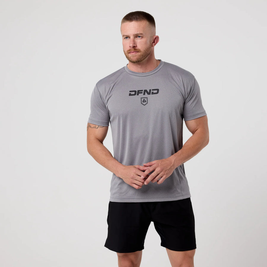 A man stands against a plain background in the DFND Speed Tech Short Sleeve + Aero Short Outdoor Kit—quick-drying gray tee and black shorts—facing forward with clasped hands, perfect for outdoor performance.