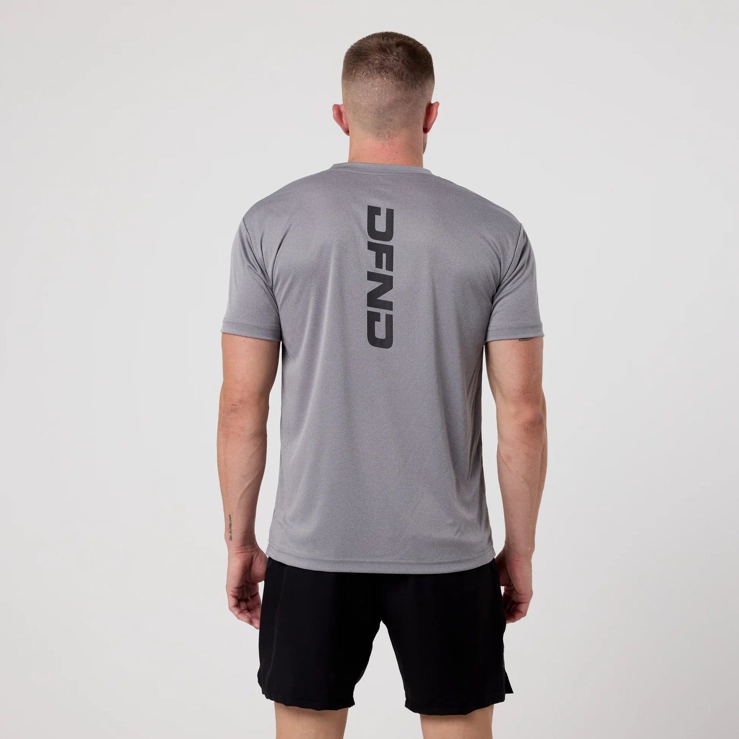 A person stands facing away, wearing the DFND Speed Tech Short Sleeve and Aero Short Outdoor Kit in gray and black, with "DFND" printed vertically on the back, made from quick-drying fabric, against a plain light background.