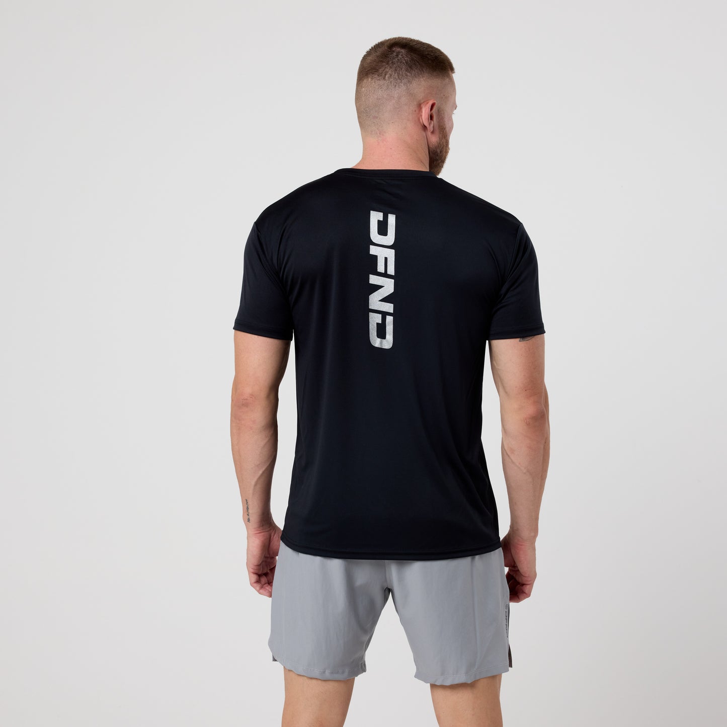 A man faces away, wearing a black DFND SPEED TECH DFND LOGO SS SHIRT and gray athletic shorts, set against a plain white background.