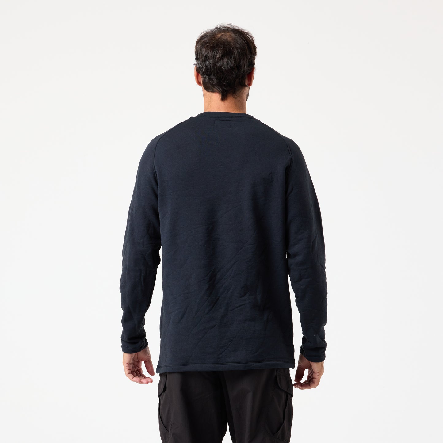 A man in a long-sleeve black DFND Shasta Fleece Crew and dark pants faces away from the camera against a white background, showcasing the top’s odor-resistant comfort.