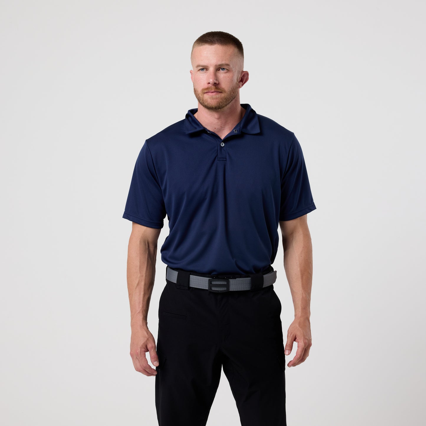 A man with short hair and a beard wears a navy blue DFND GALAXY PERFORMANCE POLO with moisture-wicking fabric and black pants, standing against a plain light background.