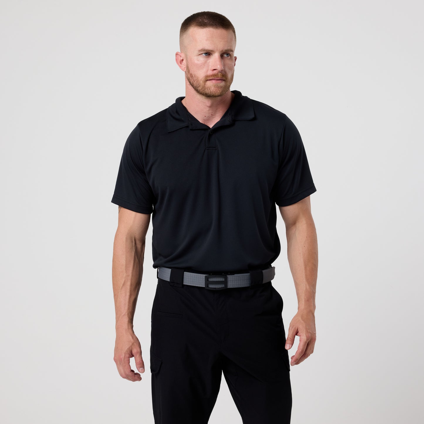 A man with short hair and a beard stands facing forward against a plain background, wearing the DFND GALAXY PERFORMANCE POLO in black, paired with black pants and a gray belt.