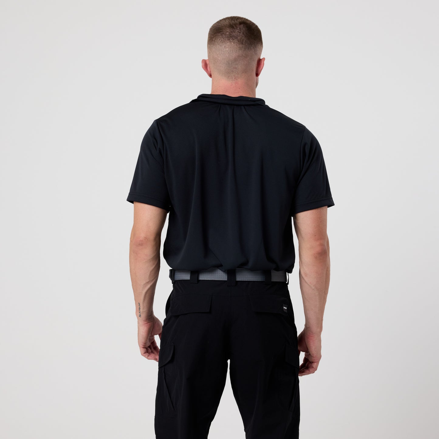 A man stands with his back to the camera wearing the DFND GALAXY PERFORMANCE POLO, a black short-sleeve shirt, paired with black pants against a plain white background.