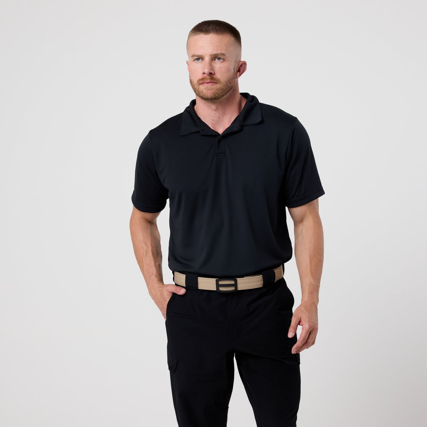 A man with short hair and a beard wears the DFND GALAXY PERFORMANCE POLO with moisture wicking, black pants, and a beige tactical belt, standing against a plain light background.