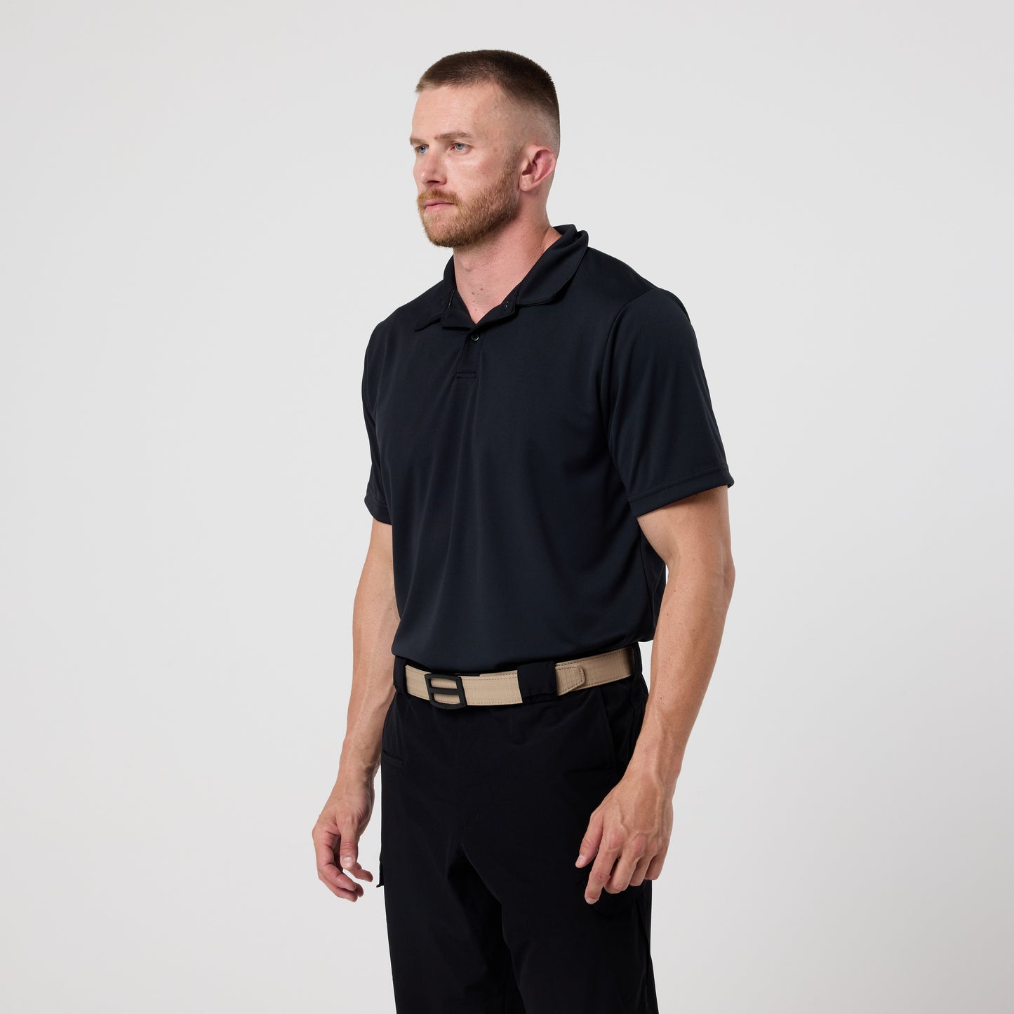 A man with short hair and a beard wears the DFND GALAXY PERFORMANCE POLO in black, paired with black pants and a beige belt, standing against a plain white background.