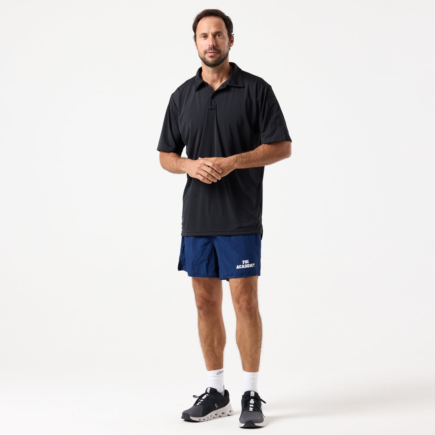 A man stands against a plain white background wearing a black polo shirt, DFND MALIBU 5" PT SHORTS FBI ACADEMY, white socks, and black sneakers.