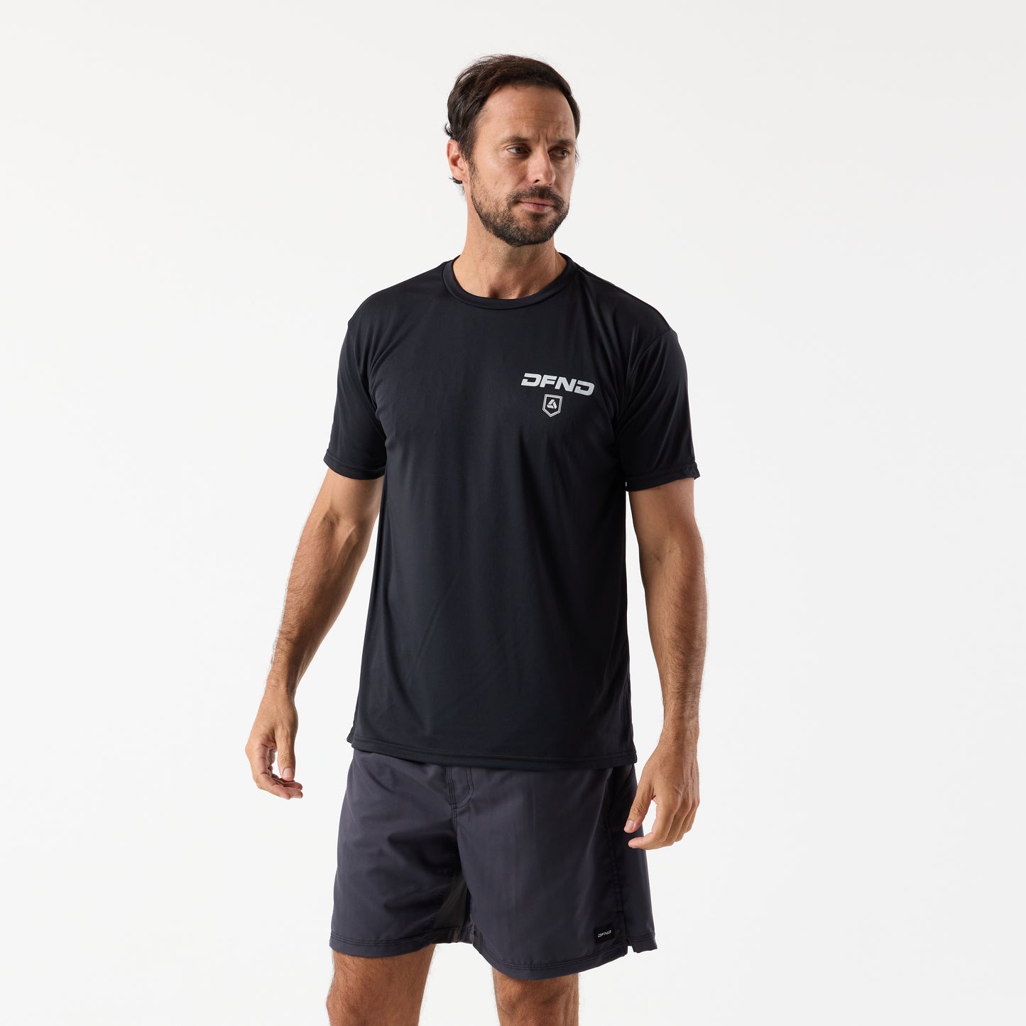A man stands against a plain white background wearing a black DFND SKULL SPEED TECH SS SHIRT - Set in Sleeve, known for its moisture-wicking design, and dark shorts, looking slightly to his left.