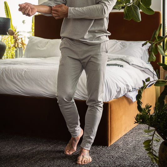 Barefoot beside a bed in a modern, plant-filled bedroom, a person adjusts their sleeve while wearing the DFND Cooling Recovery Sleepwear Long John—a light gray long-sleeve top and pants set by DFND. Infused with CELLIANT minerals to promote restful sleep, this soft infrared recovery sleepwear is thoughtfully designed for ultimate comfort in a tranquil setting.