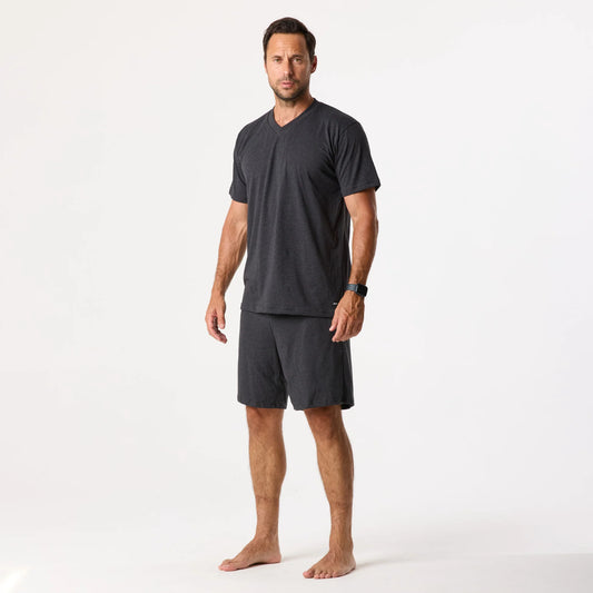 A man stands barefoot against a white background wearing DFND Sleepwear SS Shirt & Shorts, a dark gray short-sleeve V-neck set crafted with CELLIANT technology for recovery.