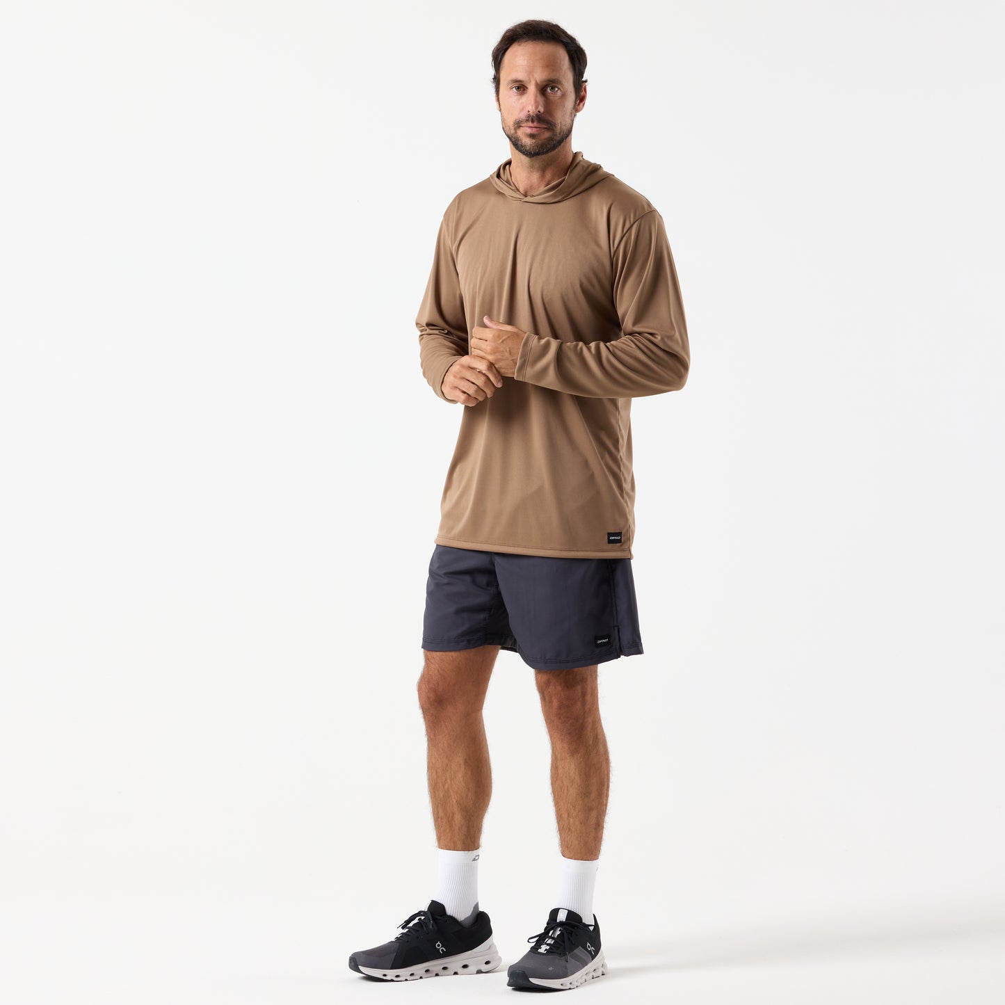 A man stands against a plain white background wearing a tan DFND SPEED TECH LS HOODED SHIRT made from antimicrobial fabric, paired with black shorts, white socks, and black athletic shoes.