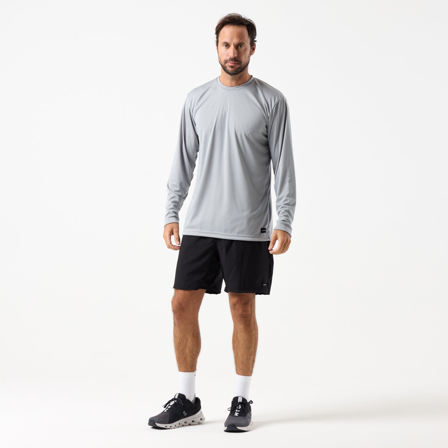 A man faces forward wearing a light gray DFND SPEED TECH LS SHIRT - Set in Sleeve, black shorts, white socks, and black athletic shoes against a plain white background.