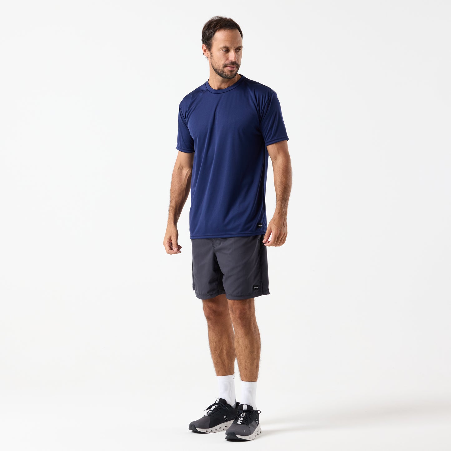 A man stands against a plain white background wearing the DFND SPEED TECH SS SHIRT - Set in Sleeve in navy, dark gray shorts, white socks, and black running shoes.