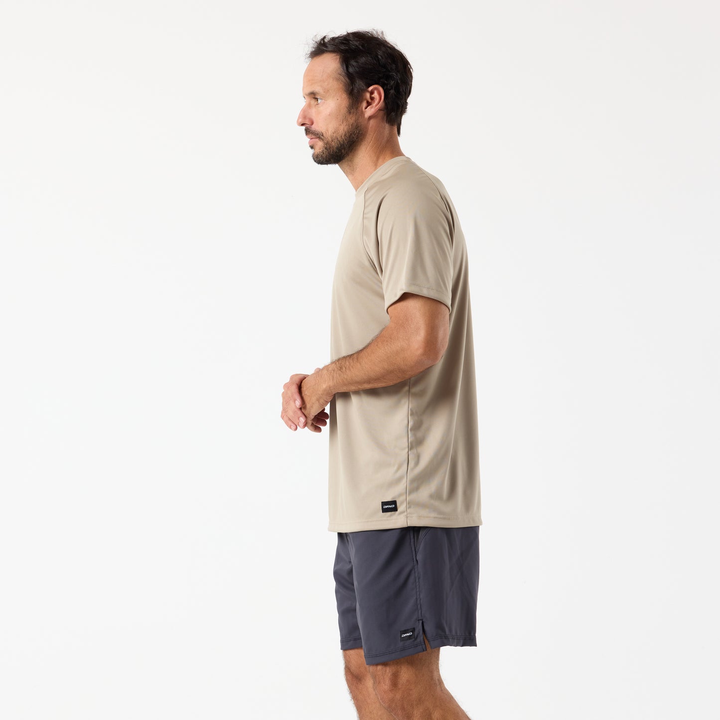 A man in a beige DFND SPEED TECH SS SHIRT - Raglan Sleeve and dark gray shorts stands in profile against a plain white background, highlighting the shirt's moisture-wicking and antimicrobial fabric.