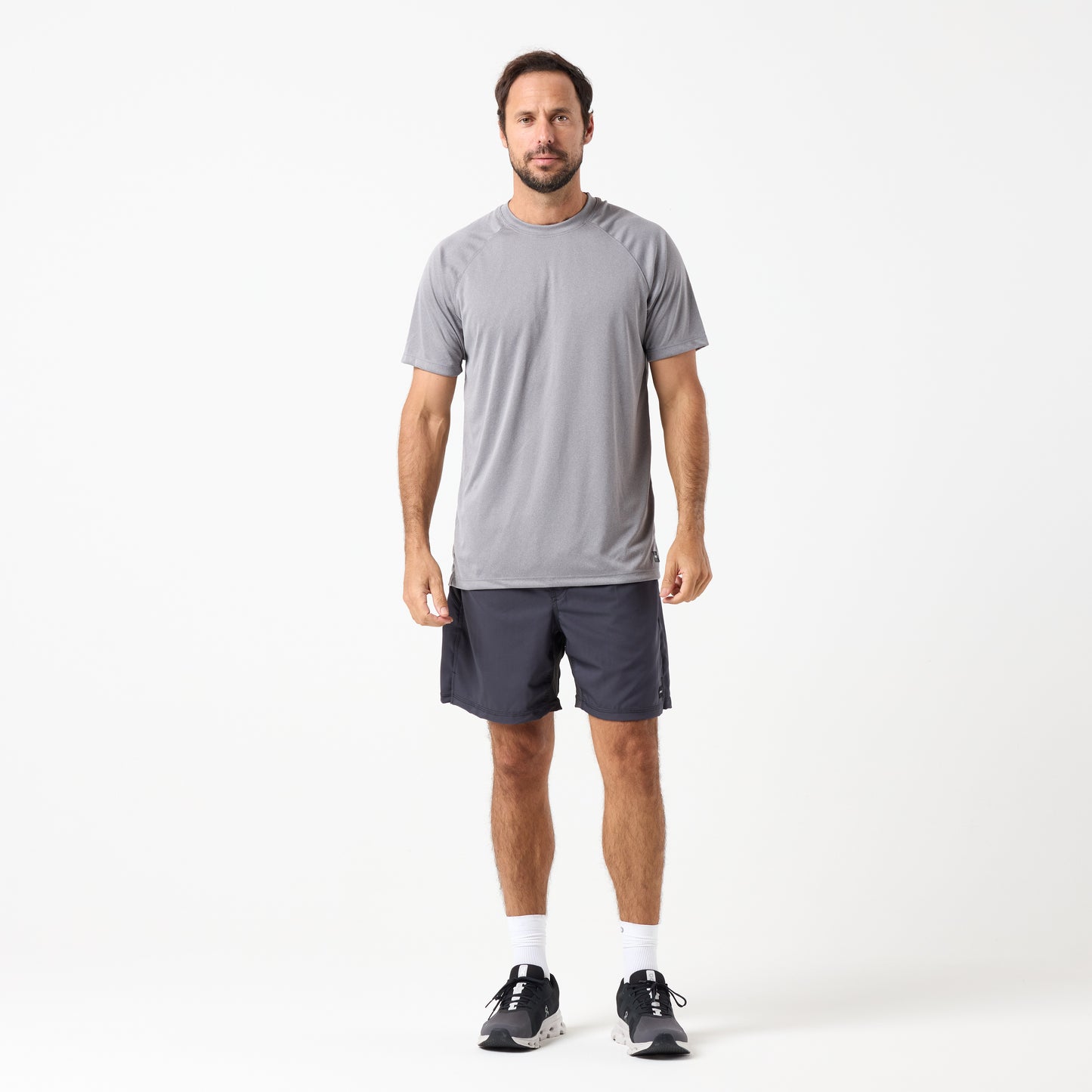 A man stands facing forward in a grey DFND SPEED TECH SS SHIRT - Raglan Sleeve with moisture-wicking fabric, dark athletic shorts, white socks, and black sneakers against a plain white background.