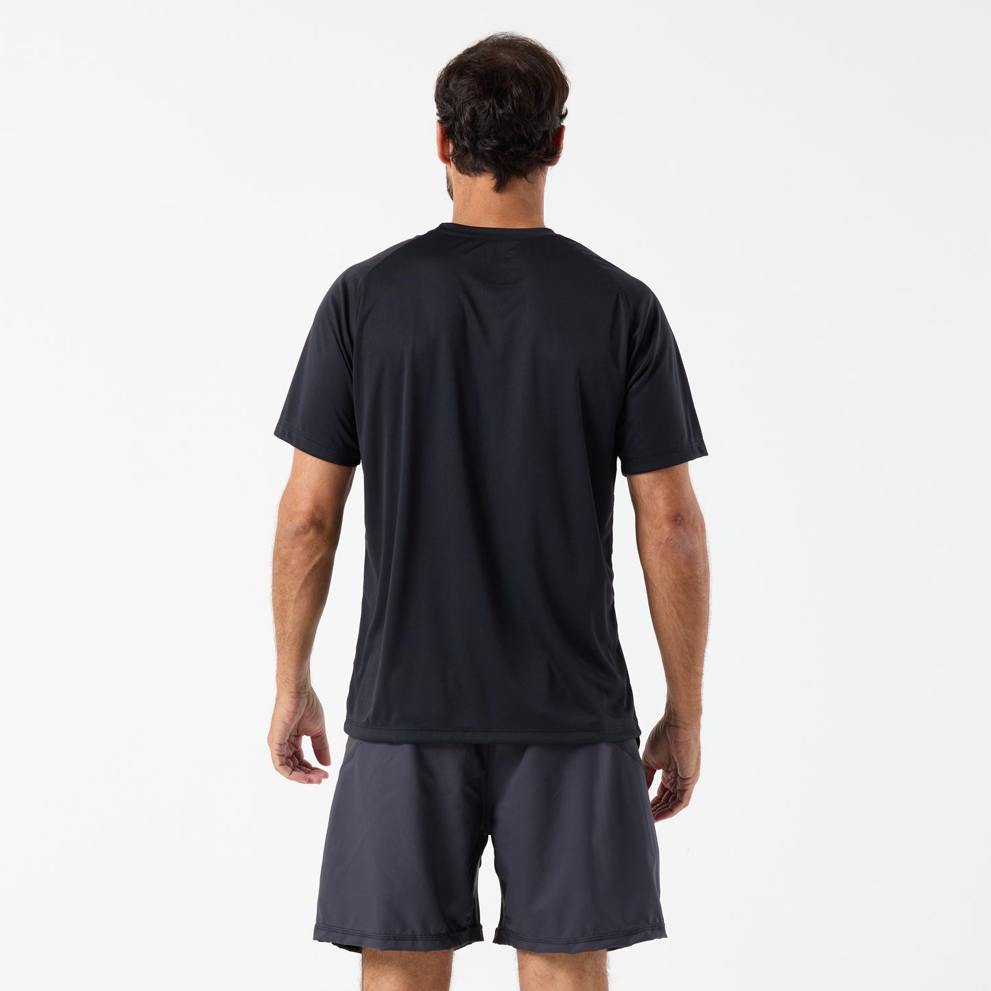 A man stands with his back to the camera, wearing a black DFND SPEED TECH SS SHIRT - Raglan Sleeve with moisture-wicking FUZE antimicrobial fabric and dark shorts, set against a plain white background.