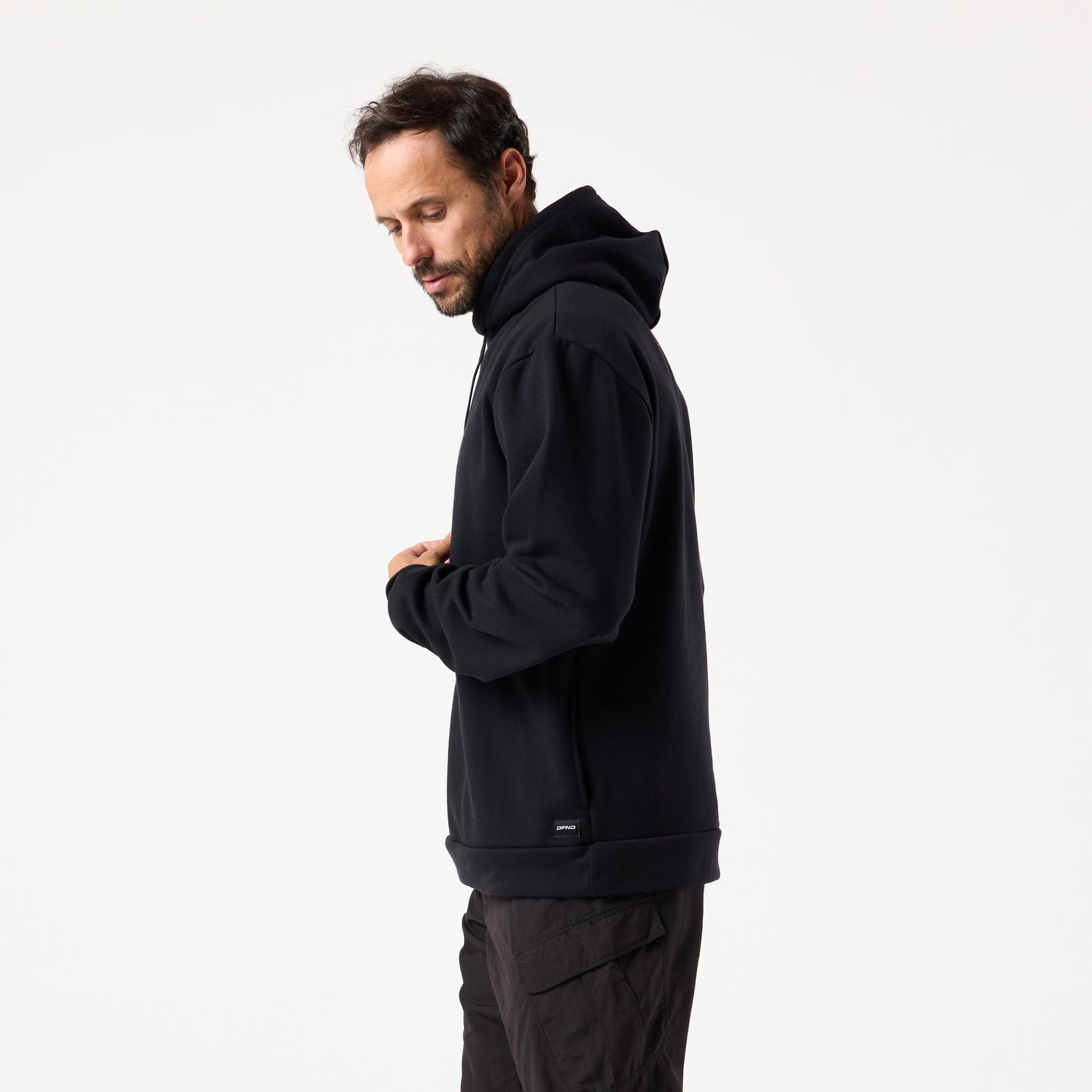 A man with short dark hair and a beard stands sideways against a plain white background, wearing the DFND TITAN FLEECE HOOD black hoodie and dark pants.