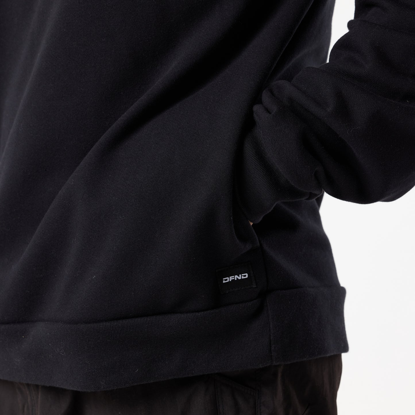 A person wears the DFND TITAN FLEECE HOOD, a black long-sleeve hoodie with a subtle label near the hem, perfect for cold weather training, standing casually with one hand in their pocket against a plain background.