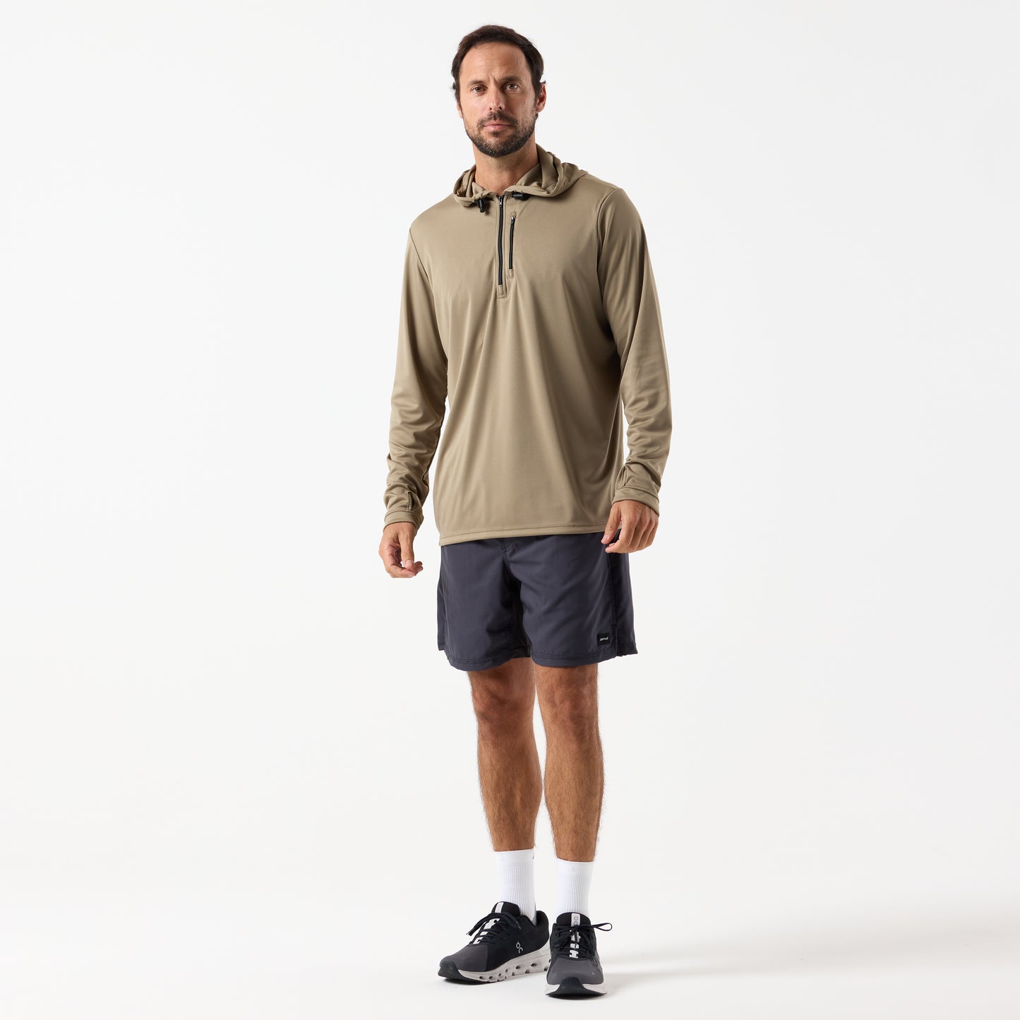 A man faces forward against a plain white background, wearing the DFND SPEED TECH HOODED 1/4 ZIP in tan, paired with dark shorts, white socks, and black athletic shoes.