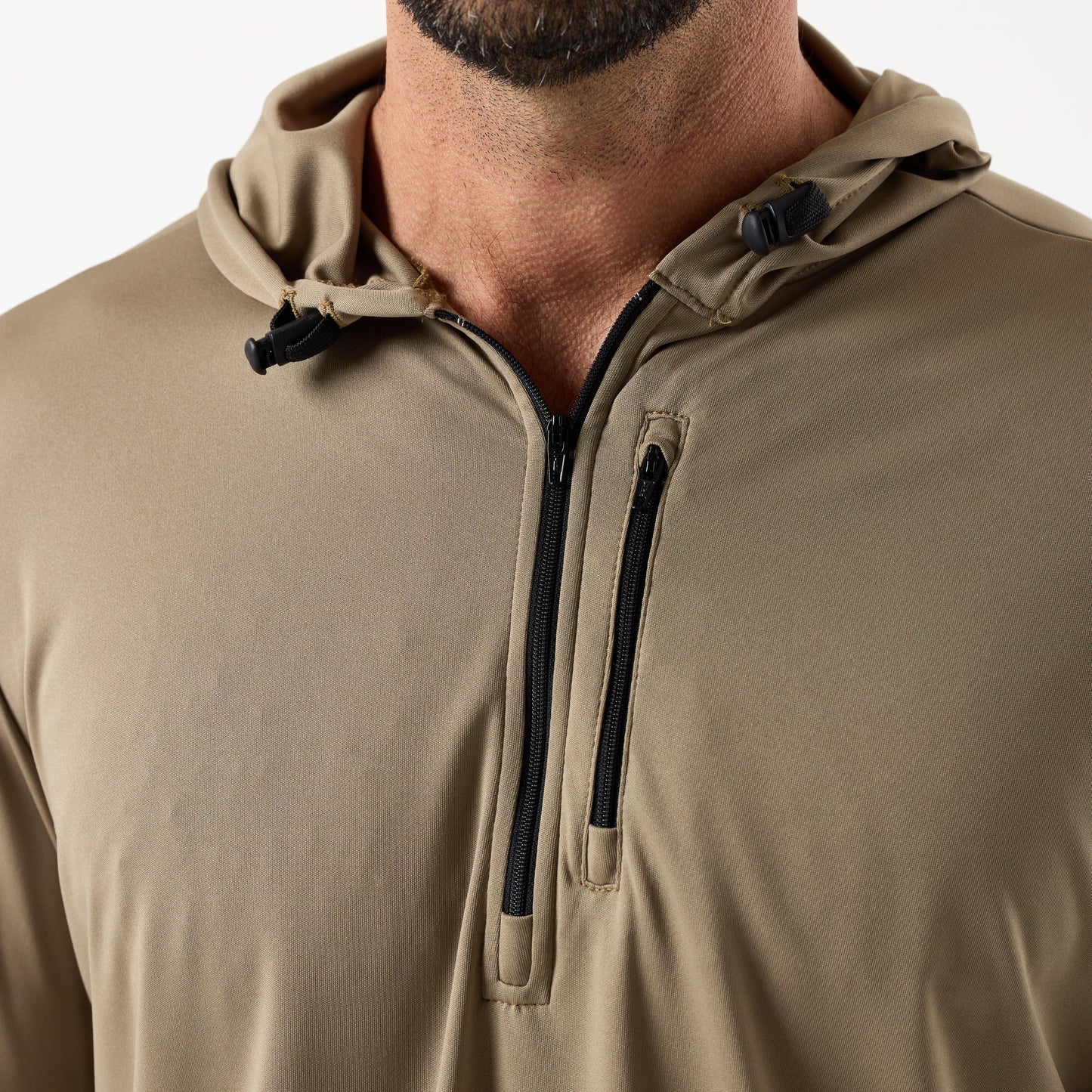 Close-up of a man wearing the DFND SPEED TECH HOODED 1/4 ZIP in tan, crafted from antimicrobial fabric with a lightweight build, half-zip front, and small zippered chest pocket.